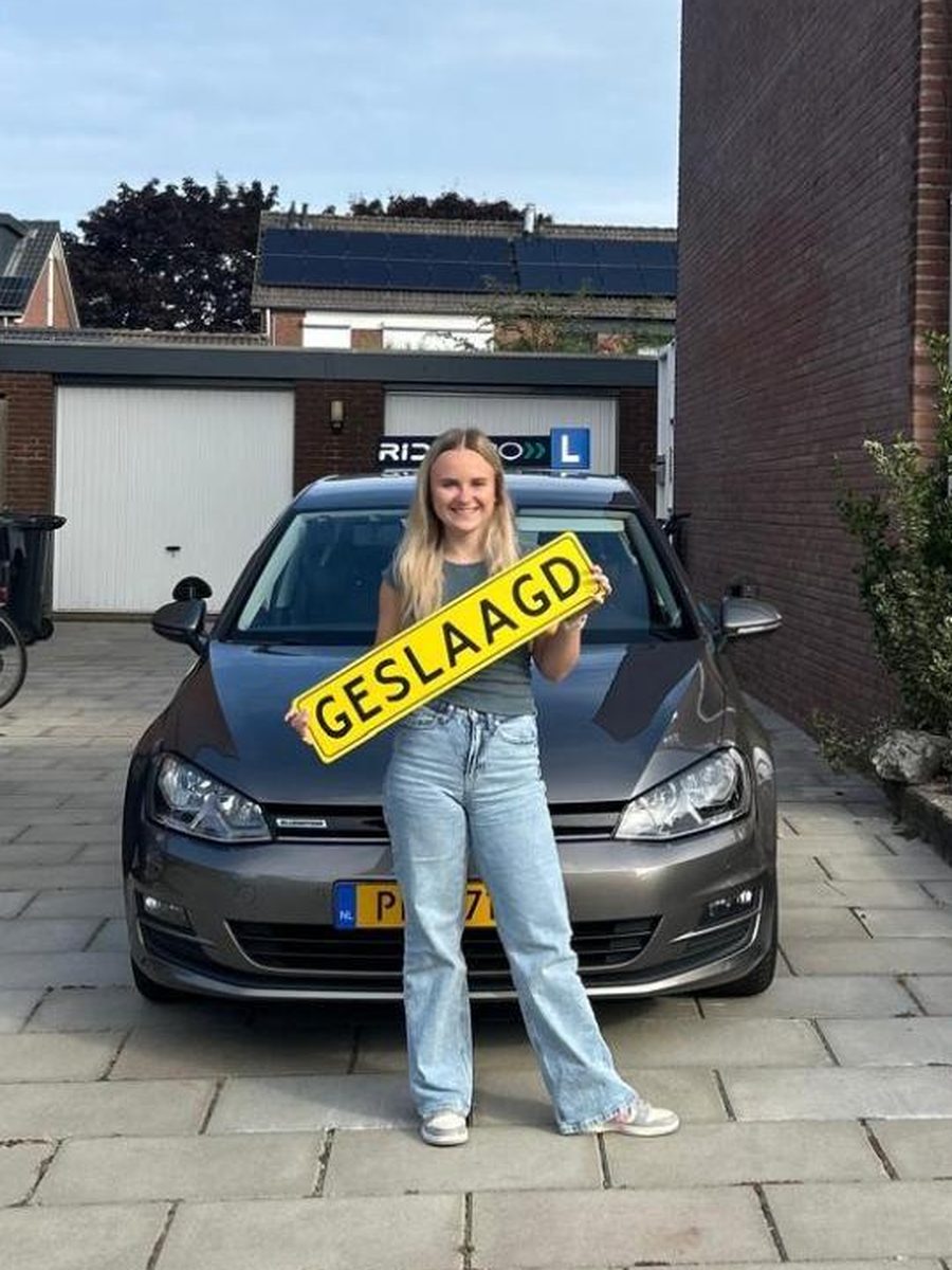 Geslaagde RidePro-leerling met het bord "geslaagd" naast de lesauto