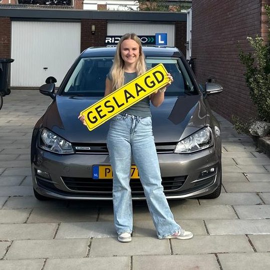 Ridepro_Geslaagd_Rijexamen_1 Blonde vrouw houdt een verkeersbord met de tekst "GESLAAGD" voor een auto.