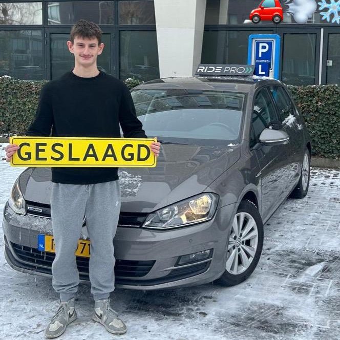Ridepro_Geslaagd_Rijexamen_2 Jonge man staat naast een grijze auto en houdt een bord met "GESLAAGD" vast.