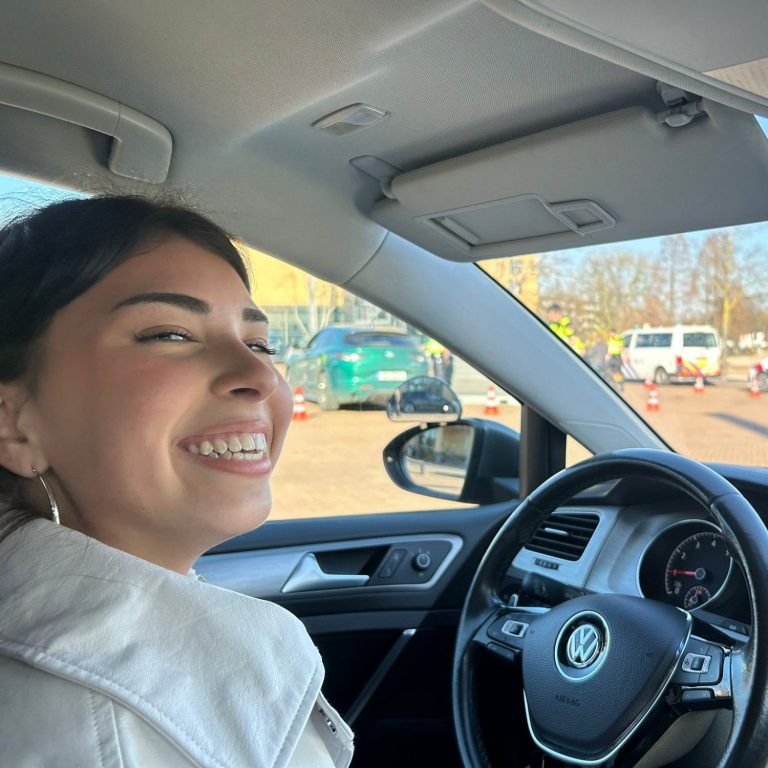 Ridepro_Rijles_Leerling_1 Jonge vrouw die vrolijk in een auto zit, met een stadsachtergrond.